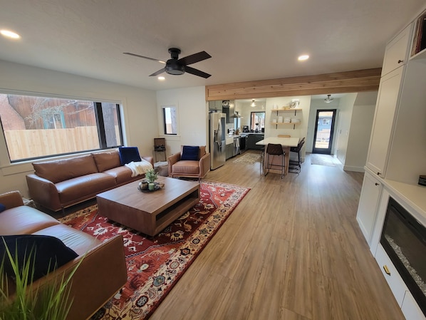 A spacious living room with a brown leather sofa, a coffee table, and a ceiling fan. There is a kitchen area with a dining table and chairs visible through an open doorway.