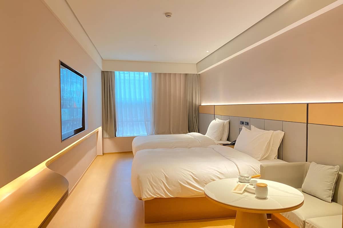 Une chambre d’hôtel avec deux lits, un canapé, une table ronde avec une tasse et une soucoupe, et une grande fenêtre avec des rideaux.