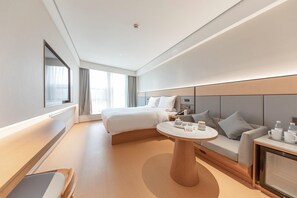 A modern hotel room with a bed, a sofa, a round table, and a bedside table with amenities.