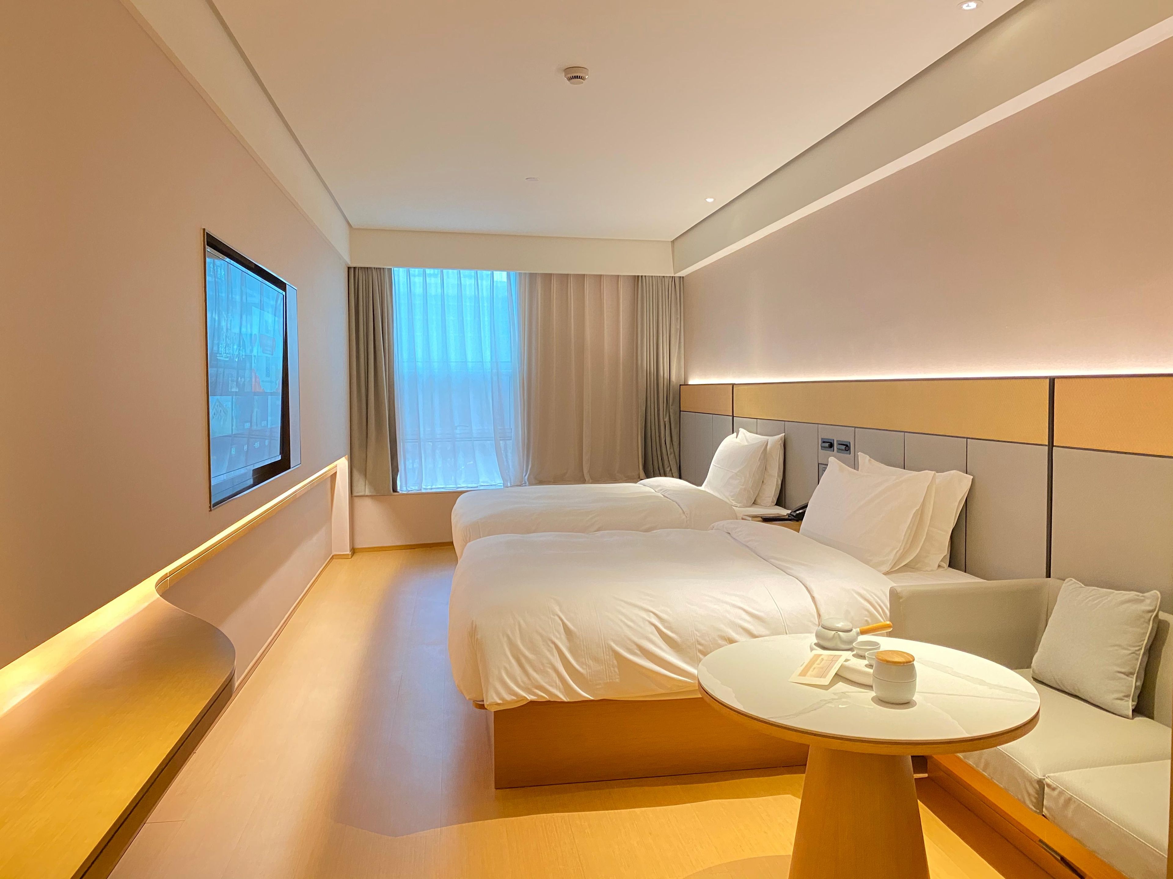 Une chambre d’hôtel avec deux lits, un canapé, une table ronde avec une tasse et une soucoupe, et une grande fenêtre avec des rideaux.