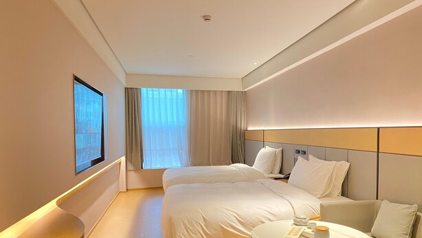 Quarto de hotel com duas camas, um sofá, uma mesa redonda com xícara e pires, e uma janela grande com cortinas.