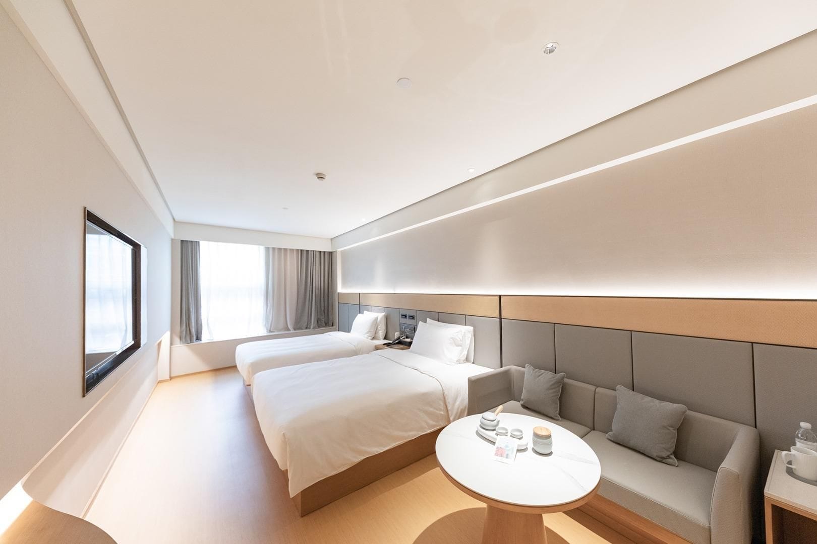A modern hotel room with two beds, a sofa, a round table, and a large window.