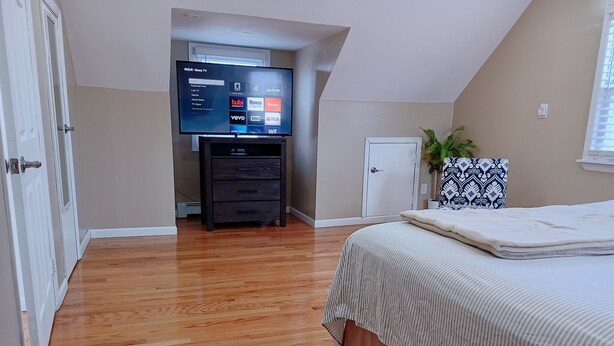 A bedroom with a wooden floor, a bed with a striped bedspread, a TV on a stand displaying streaming services, a dresser, and a door leading to another room. - Home away from home (Elmont)
