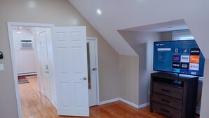 A room with a wooden floor, a white door, a TV displaying streaming services, and a dark wooden dresser. - Home away from home (Elmont)