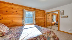 A bedroom with wooden panel walls, a large bed with a patterned bedspread, a window with curtains, and a mirror on the wall. - Beautiful mountain view! SAUNA, HOT TUB, Pool Table, and Fast WIFI! Pet friendly (Waynesville)