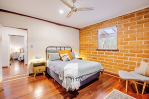 Een slaapkamer met een bakstenen muur, houten vloer, een bed met een grijze dekbedovertrek, een nachtkastje met een lamp en een raam met jaloezieën.