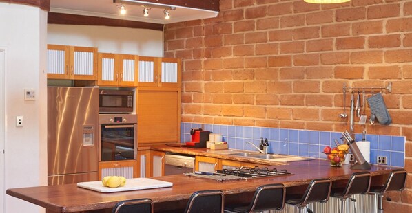 A kitchen with a brick wall, wooden cabinets, a stainless steel refrigerator, a microwave, an oven, a sink, and bar stools. - Beat the Winter Blues 25% off - XL Group and Multiple Family Getaway (Burnside Margaret River)