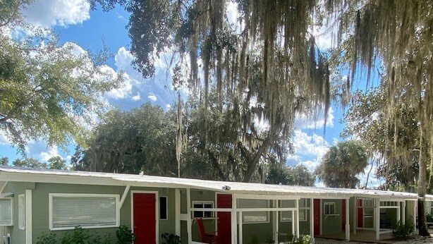A row of single-story mobile homes with white exteriors and red accents, surrounded by trees and a grassy area. - Relax by the River - Shell Harbour Room 4 on the St Johns River (Satsuma)