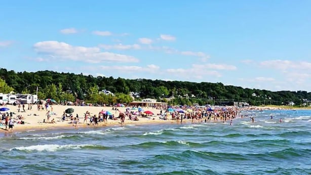 A crowded beach with people and umbrellas, a clear sky, and a forested hillside in the background.