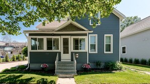 A two-story house with a front porch, a small garden with flowers, and a clear sky.