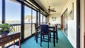 A balcony with a table and chairs, a ceiling fan, and large windows offering a view of palm trees and a body of water.