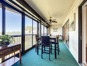 A balcony with a table and chairs, a ceiling fan, and large windows offering a view of palm trees and a body of water.