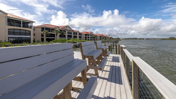 A wooden deck with benches overlooking a body of water, adjacent to a building with multiple balconies and a red roof.