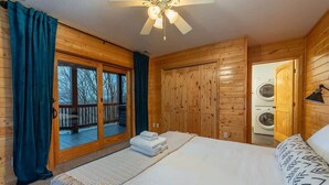 A wooden cabin bedroom with a bed, a ceiling fan, a large window with curtains, and a washing machine in the corner.
