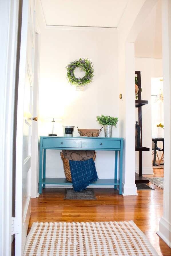 A hallway with a teal desk, a wicker basket, a blue cushion, a potted plant, and a wreath on the wall.