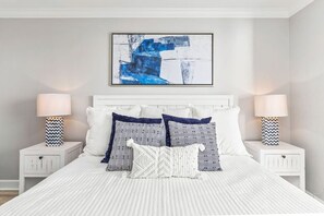 A bedroom with a white bed, two nightstands, two lamps, and a framed abstract painting above the bed.