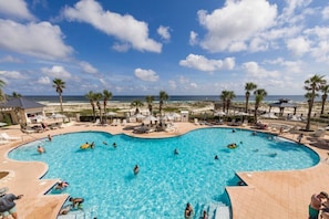 A large outdoor swimming pool surrounded by lounge chairs and umbrellas, with a beach and palm trees in the background.