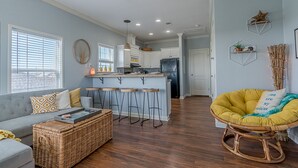 Un salon moderne comprenant un canapé, une table basse en osier, un fauteuil à bascule jaune et un coin cuisine avec un tabouret de bar.