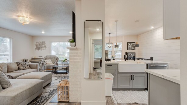 A modern living room with a sectional sofa, a coffee table, and a fireplace. There is a kitchen with a white countertop and gray cabinets.