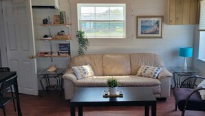 A cozy living room with a beige sofa, a coffee table, and a chair. There is a bookshelf with books and a framed picture on the wall. - Beach vibes (Pointe-du-Chêne)