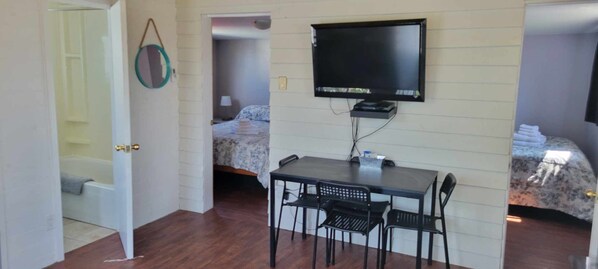 A room with a bed, a desk with chairs, a television, and a door leading to another room.