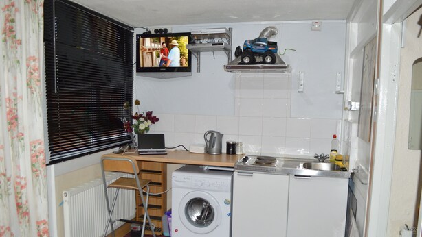 Une cuisine compacte équipée d’un lave-linge, d’un petit évier et d’un téléviseur fixé au-dessus du plan de travail.