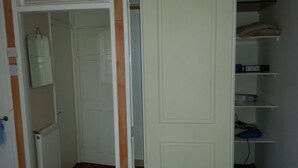 Un couloir étroit avec une porte blanche, un radiateur et une étagère sur laquelle se trouvent un coussin et une boîte.