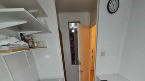 Une chambre de taille modeste, comprenant un lit, un petit bureau, une étagère avec des articles de toilette et une horloge murale.