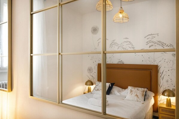 Une chambre avec une grande fenĂȘtre, un lit recouvert de draps blancs, deux lampes de chevet et un mur recouvert dâun papier peint Ă motifs floraux.