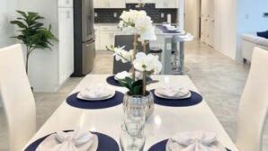 A modern dining area with a white table set for four, a vase of white orchids, and a chandelier with three lights. - Cozy 2BD/2BA Condo Near Disney 270 (Kissimmee)