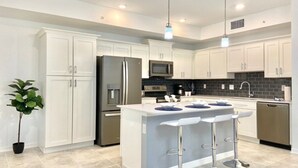 A modern kitchen with white cabinets, stainless steel appliances, and a central island with bar stools. - Cozy 2BD/2BA Condo Near Disney 270 (Kissimmee)