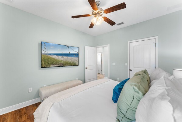 A bedroom with a large bed, a ceiling fan, a painting of a beach, and two doors.