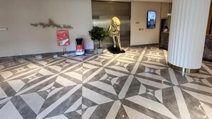 A hotel lobby with a geometric patterned floor, a reception desk, a wall-mounted digital screen, and a decorative sculpture.