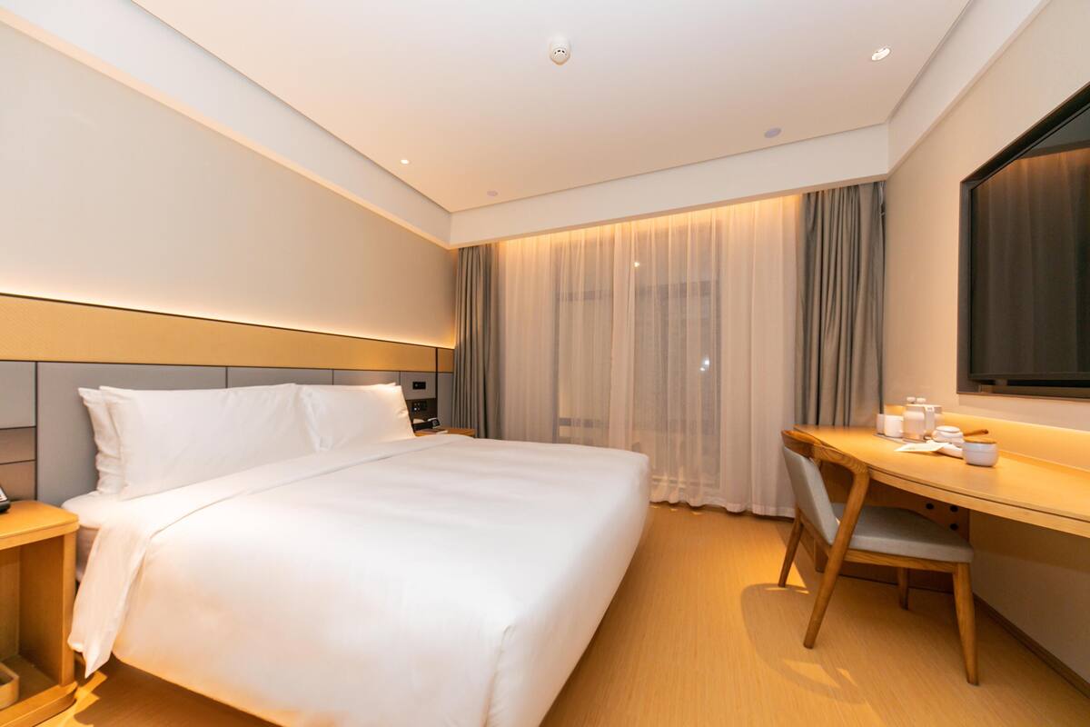 Quarto de hotel com cama, escrivaninha, cadeira, mesa de cabeceira e uma janela grande com cortinas.