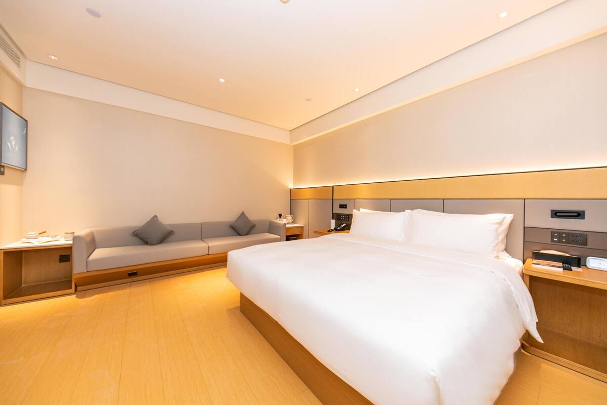 Quarto de hotel moderno com cama grande, sofá, escrivaninha e painel de controle fixado na parede.