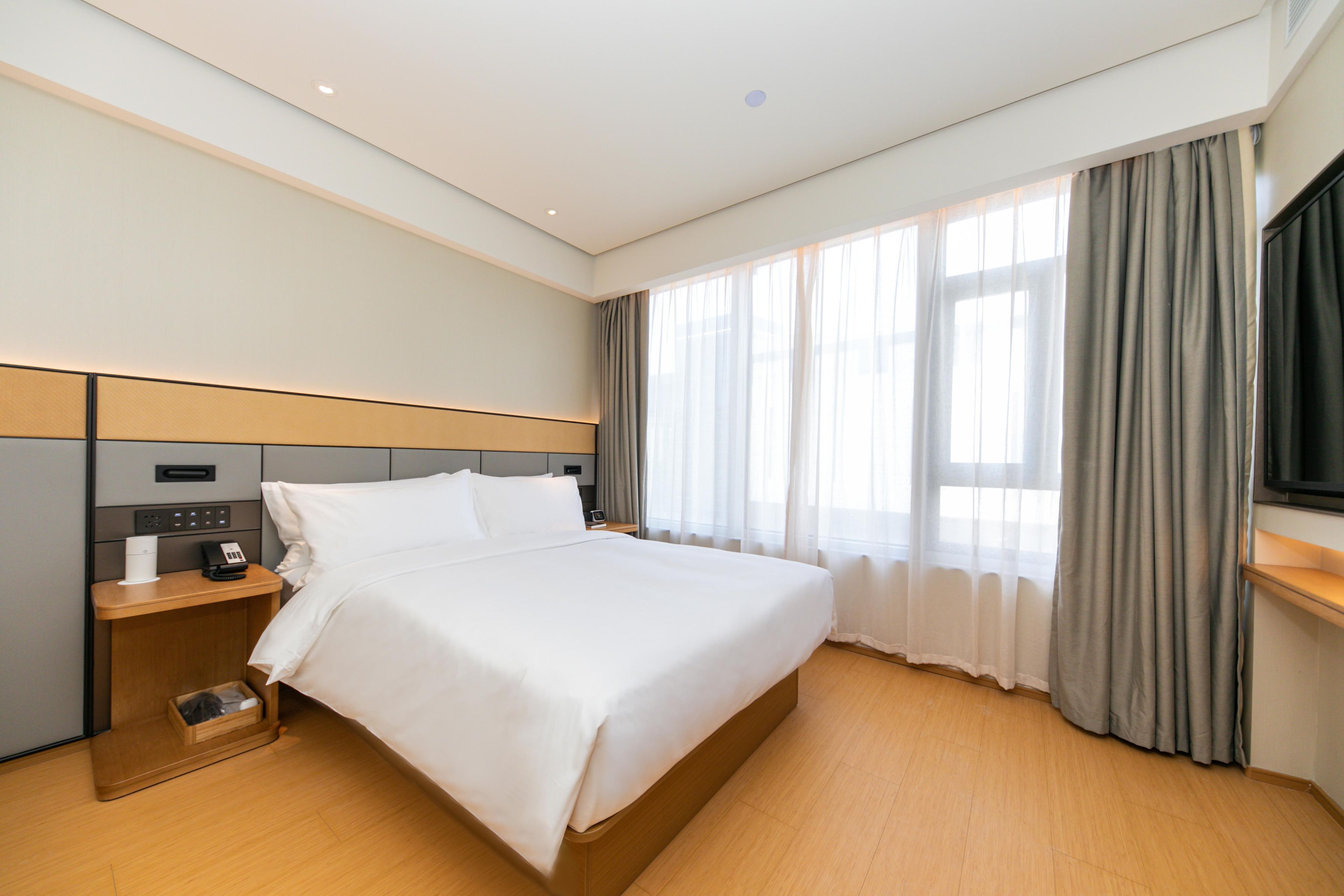 Camera d'albergo con un letto grande, un comodino con telefono e lampada, un mobiletto in legno, una TV a parete e una finestra con tende.