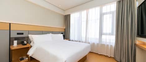 A hotel room with a large bed, a bedside table with a phone and a lamp, a wooden nightstand, a TV mounted on the wall, and a window with curtains.