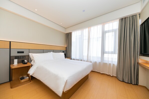 A hotel room with a large bed, a bedside table with a phone and a lamp, a wooden nightstand, a TV mounted on the wall, and a window with curtains.