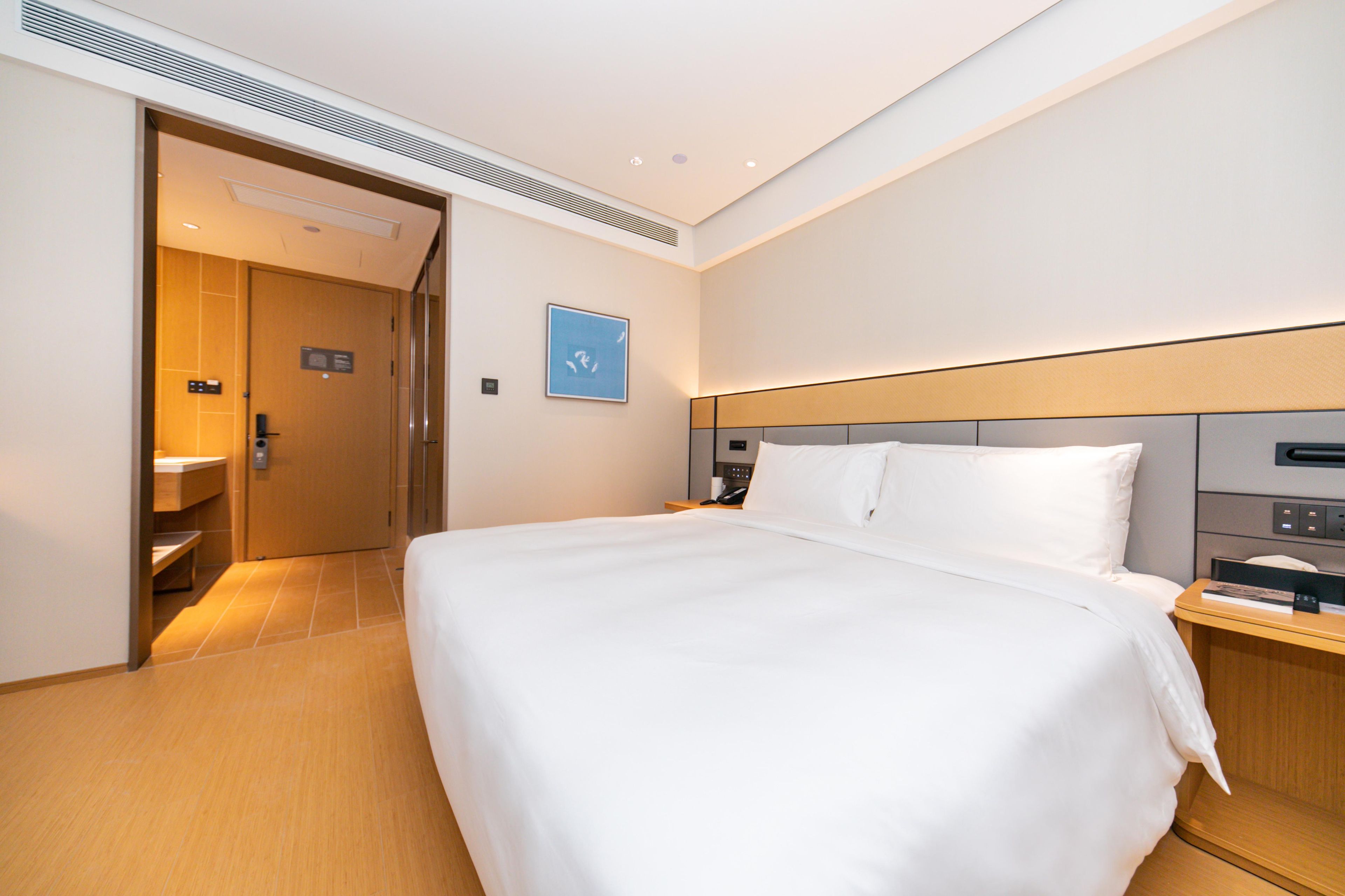 Quarto de hotel com uma cama grande, uma mesa de cabeceira, um criado-mudo, um telefone fixado na parede e uma porta que dá para o banheiro.