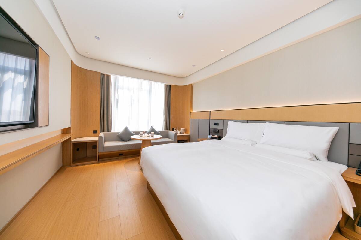 Quarto de hotel moderno com uma cama grande, área de estar com uma mesa e uma janela com cortinas.