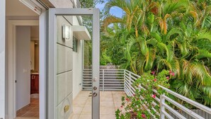 Puerta de vidrio que da a un espacio exterior cubierto con barandilla blanca, rodeado de exuberante follaje y una maceta con flores rosadas.