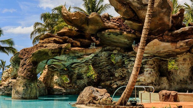 Un área de piscina con una gran formación rocosa, palmeras y una piscina azul cristalina.