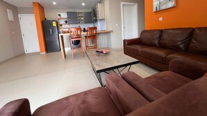 Een moderne woonkamer met een bruine leren bank, een eettafel met twee stoelen, een keuken met kasten en een koelkast, en een deur naar een andere kamer.