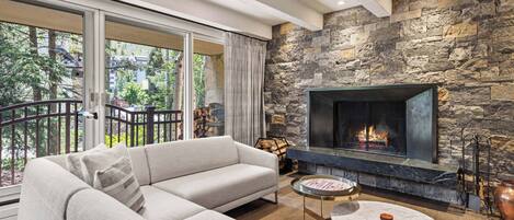 A modern living room with a stone fireplace, a large sofa, and a balcony with a view of trees and buildings.