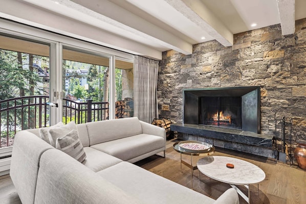 A modern living room with a stone fireplace, a large sofa, and a balcony with a view of trees and buildings. - Village Center C2J - 2 Bedroom Residence (vail)