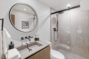 A modern bathroom with a round mirror, a sink with a marble countertop, a toilet, and a glass-enclosed shower. - Village Center C2J - 2 Bedroom Residence (vail)