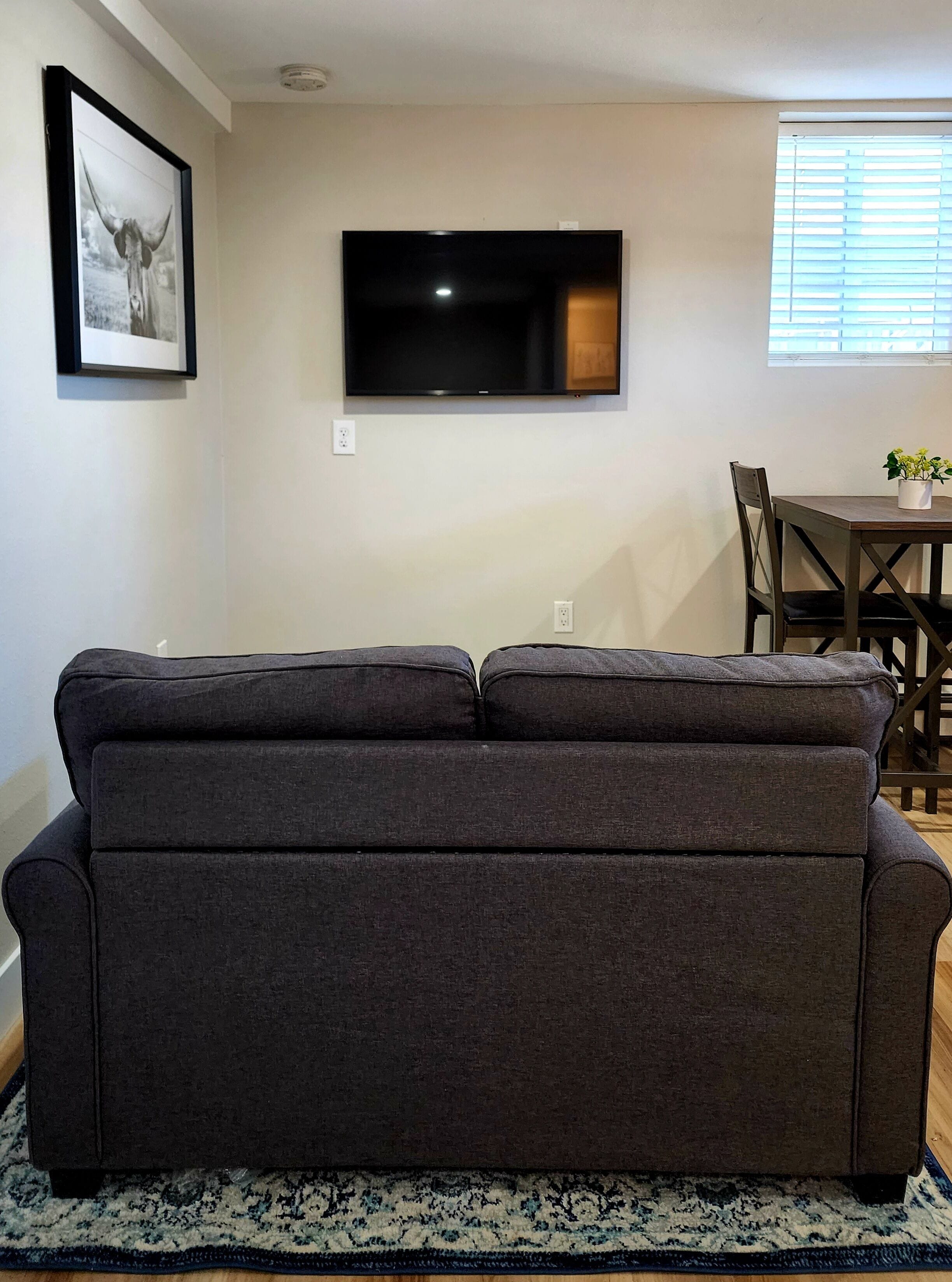 A living room with a dark grey sofa, a flat-screen TV mounted on the wall, a dining table with chairs, and a window with blinds.