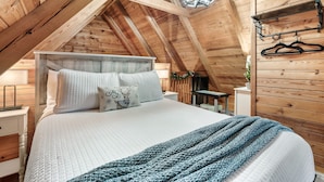 Ein gemütliches Schlafzimmer mit Holzbalkendecke, einem Bett mit weißer Bettwäsche, einer blauen Decke und einer Pendelleuchte.