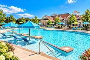 Ein Schwimmbecken mit Liegestühlen und Sonnenschirmen, umgeben von einer angelegten Grünfläche mit Bäumen und einem Gebäude im Hintergrund.