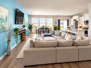 A modern living room with a large beige sectional sofa, a dining area with a table and chairs, and a kitchen with white cabinets and stainless steel appliances.